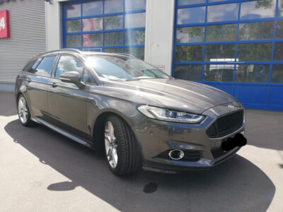 Ein gepflegter, dunkelgrauer Kombi (Ford Mondeo) steht im Sonnenlicht vor einer Werkstatt mit blauen Toren. Die Lackoberfläche des Fahrzeugs glänzt stark und spiegelt die Umgebung klar wider – ein Hinweis auf eine professionelle Keramikversiegelung. Die Versiegelung schützt den Lack vor Witterungseinflüssen, Schmutz und UV-Strahlung und verleiht ihm eine hochglänzende, spiegelähnliche Optik.