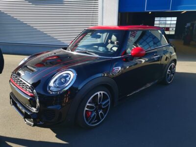 Ein schwarzer Mini Cooper mit auffälliger roter Teilfolierung an Dach, Spiegeln und Dekorstreifen auf der Motorhaube steht vor einer Werkstatt. Die glänzende Lackoberfläche deutet auf eine Keramikversiegelung hin. Die Reflexionen der Umgebung spiegeln sich klar auf dem Fahrzeug, was den hochwertigen Pflegezustand und die professionelle Versiegelung unterstreicht. Das Fahrzeug wirkt sportlich, individuell und hochwertig aufbereitet.