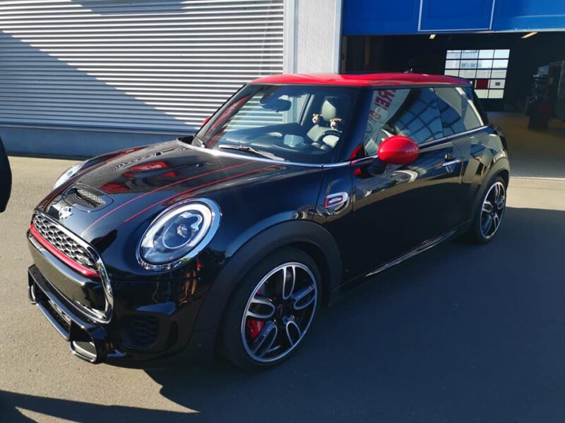 Ein schwarzer Mini Cooper mit auffälliger roter Teilfolierung an Dach, Spiegeln und Dekorstreifen auf der Motorhaube steht vor einer Werkstatt. Die glänzende Lackoberfläche deutet auf eine Keramikversiegelung hin. Die Reflexionen der Umgebung spiegeln sich klar auf dem Fahrzeug, was den hochwertigen Pflegezustand und die professionelle Versiegelung unterstreicht. Das Fahrzeug wirkt sportlich, individuell und hochwertig aufbereitet.