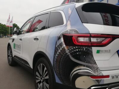 Ein weißer SUV mit auffälliger Teilfolierung parkt vor einem Gebäude. Die Folierung zeigt ein fotorealistisches Motiv des Berliner Fernsehturms, das sich von der hinteren Fahrzeugseite bis zur Tür erstreckt. Auf der Tür ist das Logo „Dietrich Dienstleistungsdienste“ samt Webadresse zu sehen. Die Gestaltung kombiniert Werbung mit urbaner Bildsprache und verdeutlicht die gestalterischen Möglichkeiten einer Fahrzeug-Teilfolierung.