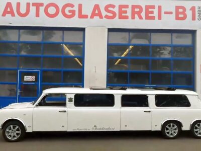 Ein weißer Stretch-Trabant mit sechs Türen steht vor dem Gebäude der Autoglaserei B1. Die überlange Karosserie des Oldtimers wurde zu einer Limousine umgebaut und trägt die Beschriftung „www.Trabi-XXL.de“ sowie „Stretchtrabant“. Das Fahrzeug wirkt außergewöhnlich und dient als Blickfang vor den blauverglasten Werkstatttoren mit dem auffälligen Schriftzug „AUTOGLASEREI-B1 GMBH“ darüber.